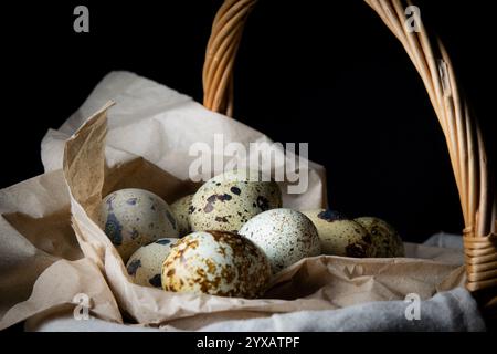 A collection of speckled quail eggs is nestled in a woven basket, resting on crumpled brown paper in low light. Stock Photo