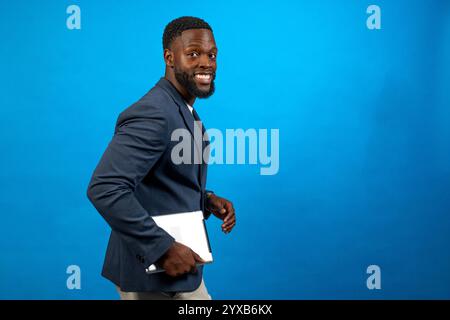 Young businessman is walking and holding a tablet, looking back over his shoulder, on a blue background Stock Photo