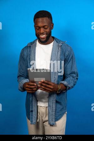 Young man reading something on a tablet, smiling, standing against a vibrant blue background Stock Photo