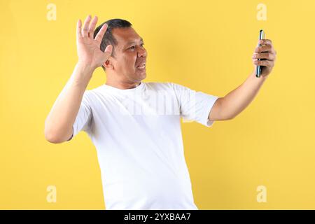Middle Age Old asian Man Wearing White tshirt Holding Mobile Phone and using it to Video Call, Isolated on Yellow Background Stock Photo