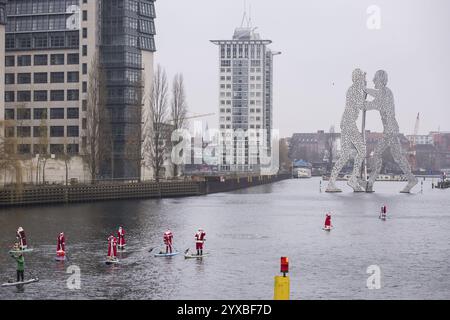 Water sports enthusiasts dressed as Santas ride SUPs on the Spree in front of the Molecule Man in Berlin on 14 December 2024 Stock Photo