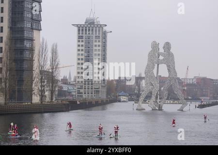 Water sports enthusiasts dressed as Santas ride SUPs on the Spree in front of the Molecule Man in Berlin on 14 December 2024 Stock Photo