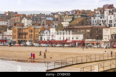 A view of a beach and town. People are on the beach and buildings line the sea front. A slipway is in the foreground and a sky with cloud is above. Stock Photo