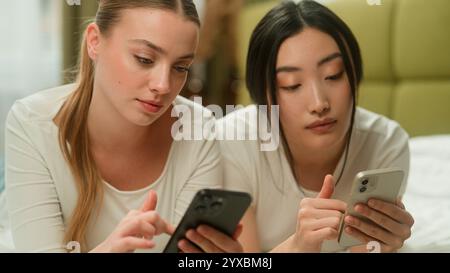 Two happy smiling multiracial friends multiethnic women diversity girls female Caucasian Asian Japanese Chinese relaxing on bed together using mobile Stock Photo