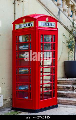 A vibrant red telephone booth was converted into a public mini library, stocked with books, and standing outdoors on a sunny day near staircases. Stock Photo