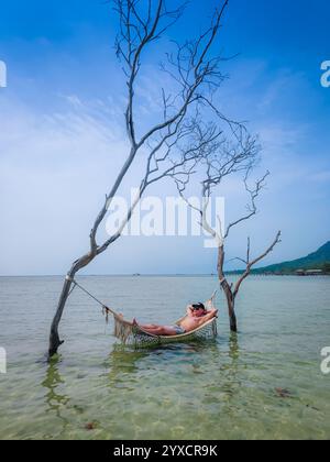 Man lying and relaxing in a hammock over water at a sea in a tropical Asian resort in Vietnam. Hammock attached to two trees in the water Stock Photo