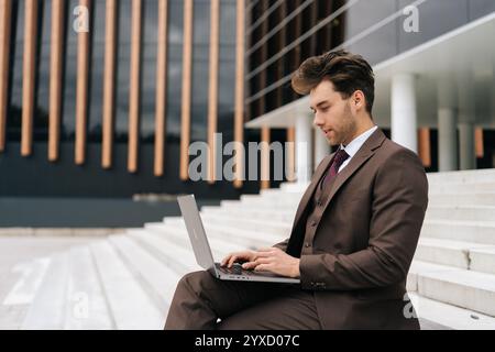 Serious ambitious young professional businessman in sleek suit diligently working on laptop computer sitting on steps by modern city office building Stock Photo