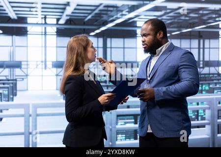 Team of engineers maintaining accurate documentation of data center systems, doing brainstorming. Teamworking colleagues talk about server room configurations and procedures, looking over clipboard Stock Photo
