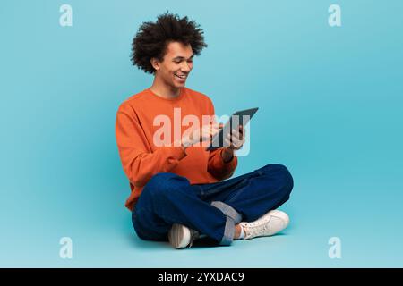 A young African American man sits cross-legged, smiling while using a tablet Stock Photo