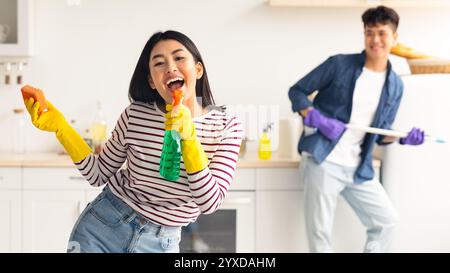 Joyful asian lady using spray as microphone, singing while cleaning Stock Photo