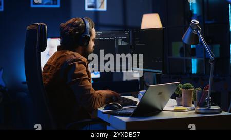 Engineer typing code in apartment office, using javascript programming language to develop software application. It admin using multimonitor desktop setup and notebook to finish tasks, camera A Stock Photo