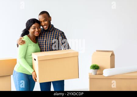 Couple Moving. Happy Black Spouses Holding Cardboard Box And Smiling At Camera Stock Photo