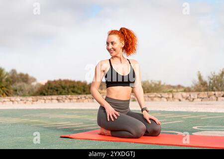 Happy sporty woman sitting in Vajrasana posture on exercise mat at outdoor gym against sky. Smiling young female in activewear is looking away while doing yoga at park during daytime.  Stock Photo