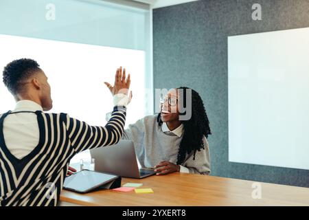 This image shows two colleagues sharing a high-five in an office setting, symbolizing teamwork and success. They appear enthusiastic and engaged, capt Stock Photo
