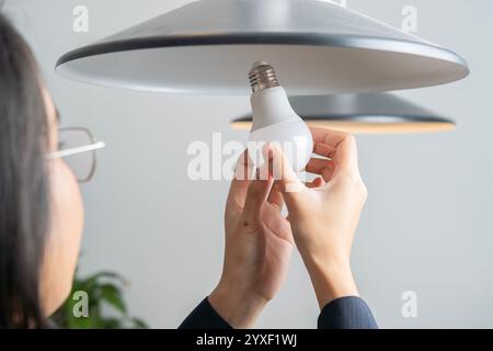 Back view shot of Asian woman changing broken light bulb to LED at home. Selective focus on hands. Stock Photo