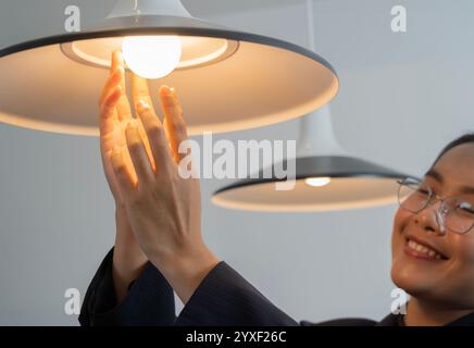 Happy young Asian woman changing a light bulb at the home office. Electricity replacing concept. Banner. Selective focus. Stock Photo
