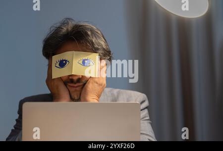 Lazy Asian male employee have stickers with painted eyes feel overwhelmed with computer work in office at night. Stock Photo