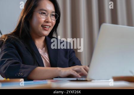 Smiling successful young Asian woman working with data e-commerce at her desk on a laptop computer. Selective focus. Stock Photo