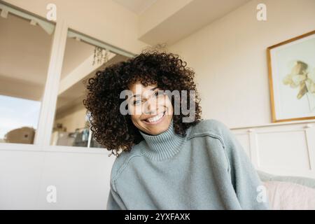 A cheerful woman with curly hair smiling warmly while sitting comfortably in a cozy bedroom. Stock Photo