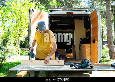 Carpenter using electric plane on wooden plank, constructing camper van outdoors Stock Photo
