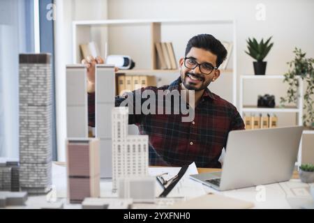Young Indian male architect working on city planning with laptop and scale models. Focused professional at work in modern office setting. Concept of architecture, creativity, urban planning. Stock Photo
