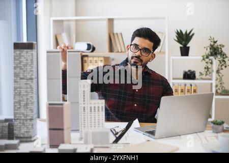 Young Indian male architect working on city planning with laptop and scale models. Focused professional at work in modern office setting. Concept of architecture, creativity, urban planning. Stock Photo
