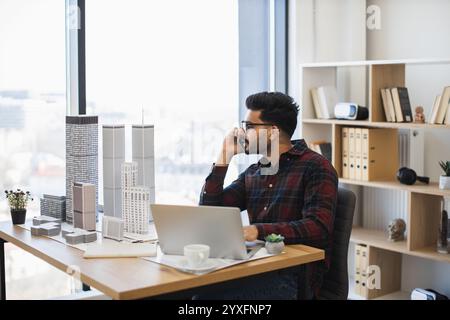 Indian male architect in office having phone call while working on modern city model. Young professional in plaid shirt focused on architectural designs by large window. Stock Photo