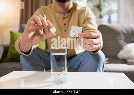 Close-up photo of young man dropping effervescent tablet into glass of water, with tablet dissolving sitting at table in living room. Stock Photo