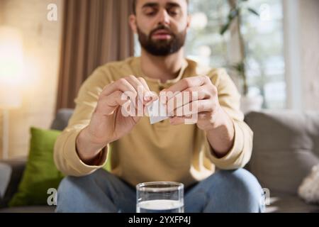 Close up of young sick man opens white sachet, preparing to take effervescent tablet while sitting at table on sofa in living room. Stock Photo