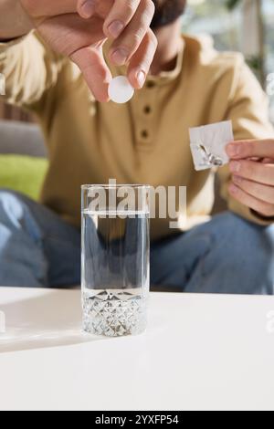 Close-up photo of young man dropping effervescent tablet into glass of water, with tablet dissolving sitting at table in living room. Stock Photo