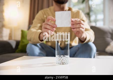 Close up of young sick man shows white sachet, preparing to take effervescent tablet while sitting at table on sofa in living room. Stock Photo