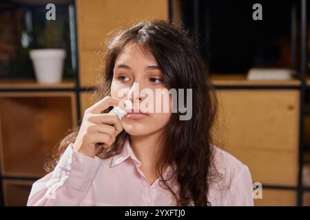 Young woman sitting at table, suffering from allergy, runny nose or nasal congestion in office and drops her nose with nasal spray Stock Photo