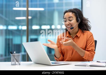 A cheerful businesswoman wearing a headset engages in a video conference using her laptop. She gestures expressively while seated in a modern office space, emphasizing connectivity and communication Stock Photo