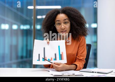 A professional woman explains data trends using graphs during a business meeting in a modern office. Her presentation focuses on statistical analysis while fostering teamwork and professional growth Stock Photo