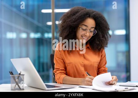Smiling woman working in a modern professional office setting, reviewing documents using laptop. The bright and contemporary environment Stock Photo