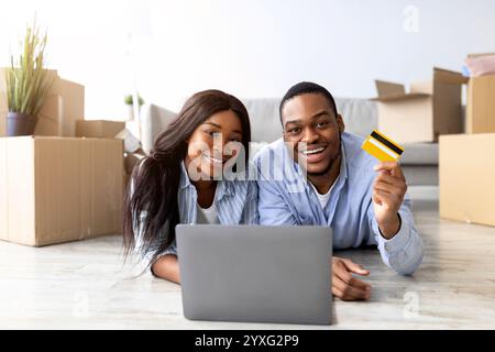 Loving black spouses buying house decor online, using laptop and credit card, lying on floor among carton boxes Stock Photo