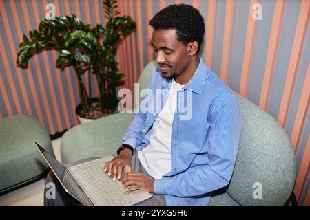 Medium shot of relaxed African American male employee in striped blue shirt typing on laptop keyboard while working on comfy office seats in coworking lounge area, camera flash, copy space Stock Photo