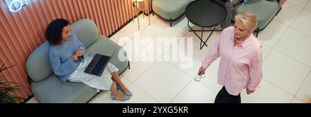 Website header shot of multiethnic woman video calling while sitting on comfy couch in coworking lounge area while working with other employees in common area Stock Photo