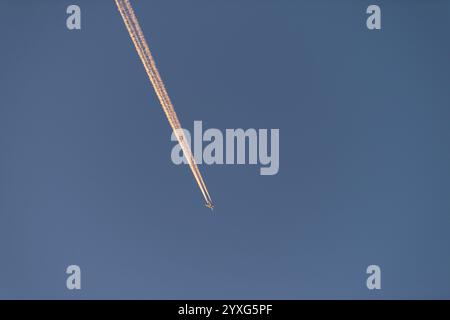 Passenger jet plane flying high in distant clear sky, leaving smoke contrail catching the golden light from the sunrise. Stock Photo