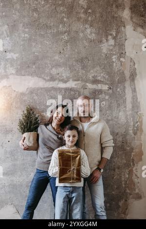 Family with a small Christmas tree and a present standing in front of a concrete wall Stock Photo