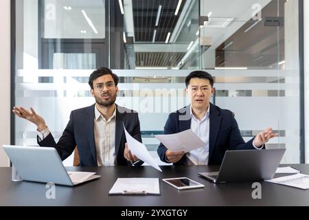 Two business professionals sitting in a modern office, visibly expressing disappointment and frustration. They are holding documents, showcasing a tense and difficult situation. Stock Photo