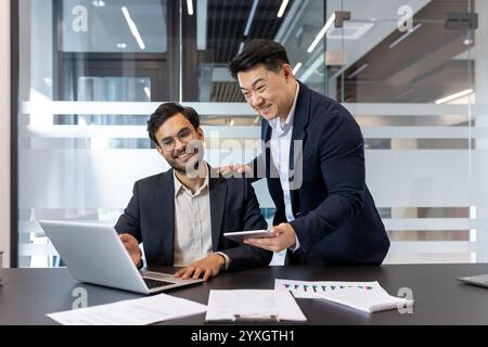 Two colleagues work together in a sleek office environment. They appear engaged and cooperative, using a laptop and a tablet to discuss or oversee tasks. Stock Photo