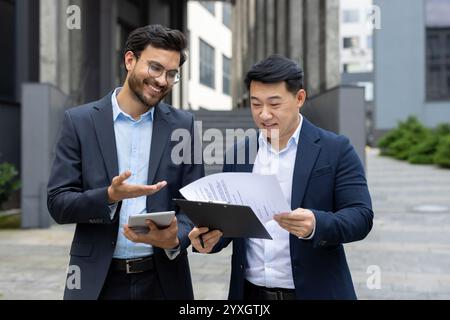 Two business professionals collaborating reviewing documents outdoors. One holds a tablet while they discuss details regarding a clipboard with printed papers. They appear engaged and happy working Stock Photo