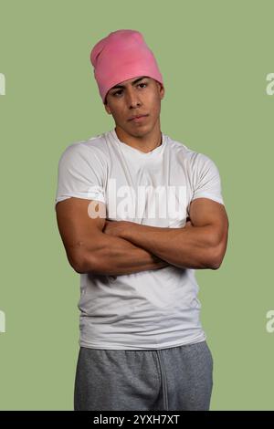 Studio portrait of a serious young latino man wearing pink beanie and white t-shirt, crossing his arms on green background Stock Photo