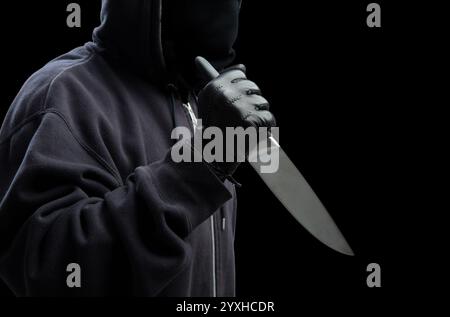 Criminal holding a knife, hooded man holding a weapon in gloved hand, black background, armed attack concept. Stock Photo