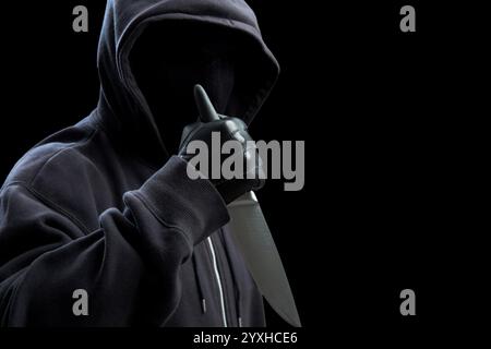 Criminal holding a knife, hooded man holding a weapon in gloved hand, black background, armed attack concept. Stock Photo