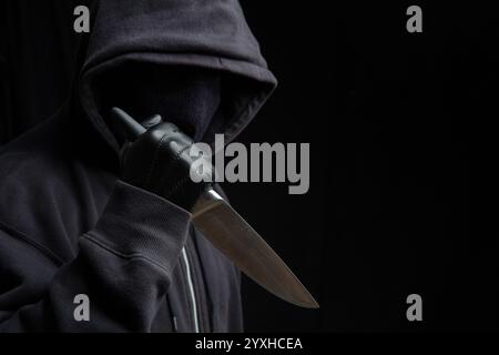Criminal holding a knife, hooded man holding a weapon in gloved hand, black background, armed attack concept. Stock Photo