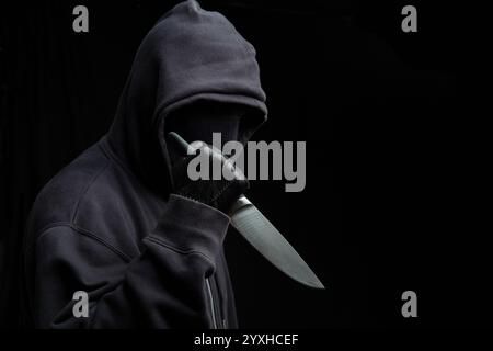 Criminal holding a knife, hooded man holding a weapon in gloved hand, black background, armed attack concept. Stock Photo