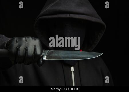 Criminal holding a knife, hooded man holding a weapon in gloved hand, black background, armed attack concept. Stock Photo