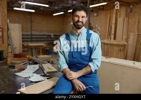 Skilled young adult woodworker sitting on workbench in well-equipped workshop Stock Photo